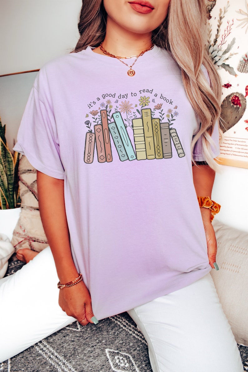 K&ouml;nnte beinhalten: Helllila T-Shirt mit dem Text "it's a good day to read a book" &uuml;ber einer Grafik von B&uuml;chern und Blumen. Das Shirt hat eine lockere Passform und einen bequemen Stil.