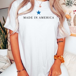 K&ouml;nnte beinhalten: Wei&szlig;es T-Shirt mit einem blauen Stern und dem Text "MADE IN AMERICA" auf der Vorderseite.