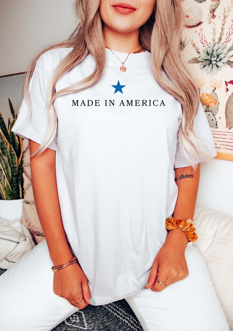 K&ouml;nnte beinhalten: Wei&szlig;es T-Shirt mit einem blauen Stern und dem Text "MADE IN AMERICA" auf der Vorderseite.