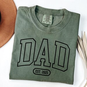May include: A green t-shirt with the word "DAD" in black block letters and the text "EST. 2025" in a black oval below.