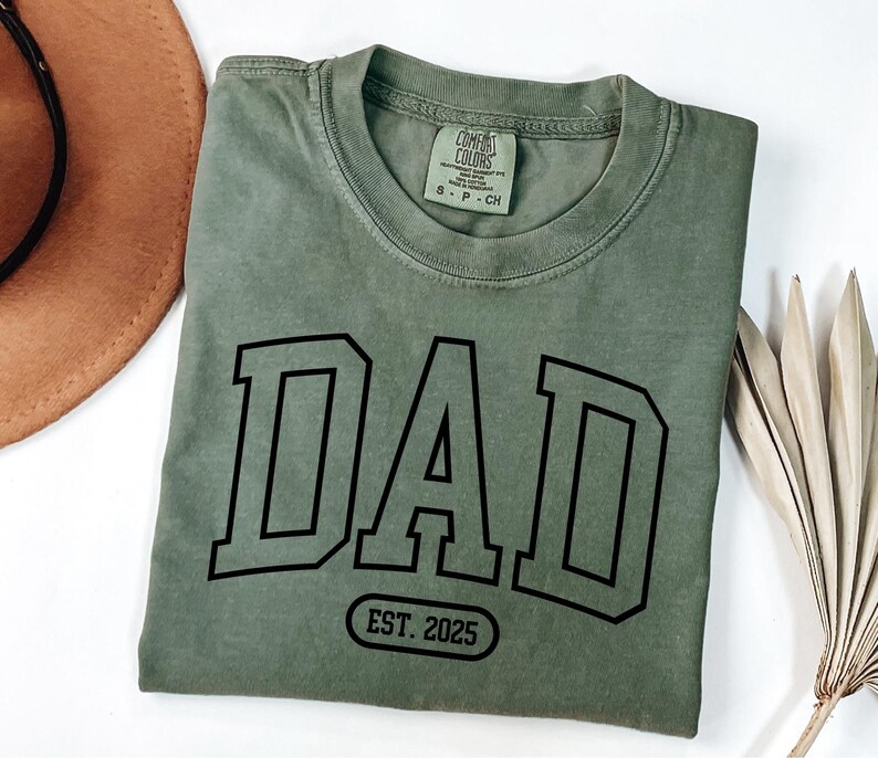 May include: A green t-shirt with the word "DAD" in black block letters and the text "EST. 2025" in a black oval below.