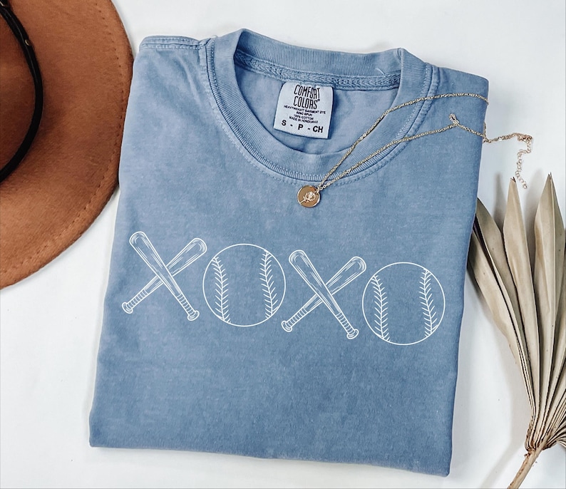 May include: A light blue t-shirt featuring a white graphic of two baseball bats crossed over each other with a baseball on either side of the bats. The graphic says "XOXO" with the baseballs forming the "O". The shirt has a tag that says "COMFORT COLORS" and "S - P - CH".