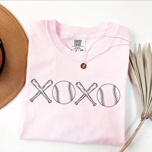 May include: Pink t-shirt featuring a graphic of two baseball bats crossed over each other with a baseball on either side of the bats. The graphic reads "XOXO".