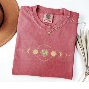 K&ouml;nnte beinhalten: Ein rotes T-Shirt mit einem goldenen himmlischen Design mit einem Globus, Monden und Sonnen. Das Shirt hat ein Comfort Colors-Etikett mit der Gr&ouml;&szlig;e S-P-CH.