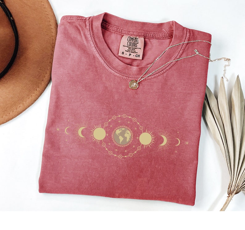 K&ouml;nnte beinhalten: Ein rotes T-Shirt mit einem goldenen himmlischen Design mit einem Globus, Monden und Sonnen. Das Shirt hat ein Comfort Colors-Etikett mit der Gr&ouml;&szlig;e S-P-CH.