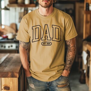 May include: A mustard yellow t-shirt with a black graphic that says "DAD EST. 2025".