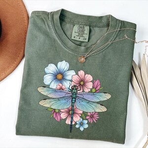 May include: Olive green Comfort Colors t-shirt featuring a colorful dragonfly design with blue, pink, and purple wings, surrounded by flowers. The shirt has a crew neck and short sleeves. A gold necklace with a pendant is on top of the shirt.