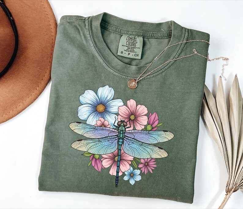 May include: Olive green Comfort Colors t-shirt featuring a colorful dragonfly design with blue, pink, and purple wings, surrounded by flowers. The shirt has a crew neck and short sleeves. A gold necklace with a pendant is on top of the shirt.