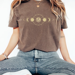K&ouml;nnte beinhalten: Ein braunes T-Shirt mit einem goldenen Aufdruck eines himmlischen Designs mit Sonne, Mond und Erde.