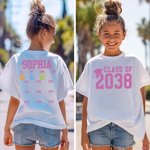May include: White t-shirt with pink text and graphics. The front of the shirt says "CLASS OF 2038" with a graduation cap. The back of the shirt says "SOPHIA" with grade levels and handprints.