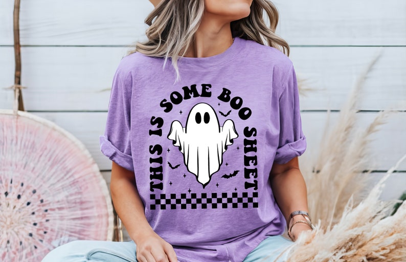 以下が含まれることがあります： 白い幽霊と黒い文字「This is some boo sheet」がプリントされた紫色のTシャツ。幽霊は2つの目があり、コウモリと星に囲まれています。