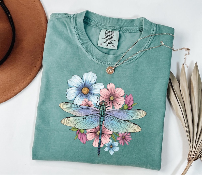 May include: A teal Comfort Colors t-shirt featuring a colorful dragonfly design with blue, pink, and yellow flowers. The shirt has a round neckline and short sleeves. A silver necklace with a small pendant is also visible.