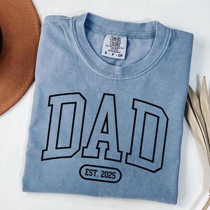 May include: A light blue t-shirt with a black graphic that says "DAD EST. 2025" in a varsity style font.