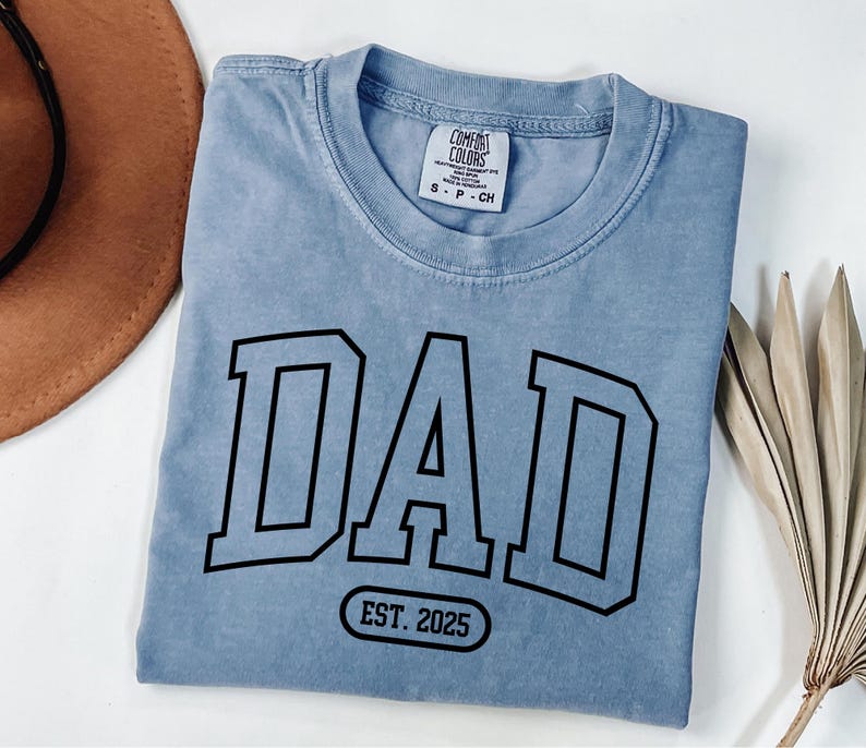 May include: A light blue t-shirt with a black graphic that says "DAD EST. 2025" in a varsity style font.