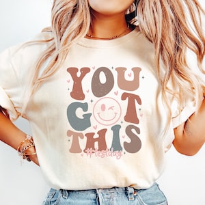 May include: A white t-shirt with a graphic design that says "You Got This" in a retro font with a winking smiley face and the hashtag "#Yestoday".