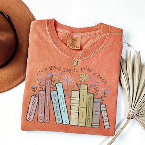 K&ouml;nnte beinhalten: Orangefarbenes Comfort Colors T-Shirt mit dem Schriftzug "it's a good day to read a book" und einer Grafik von B&uuml;chern und Blumen. Eine goldene Halskette und ein brauner Hut sind ebenfalls im Bild.