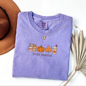 May include: A lilac Comfort Colors t-shirt featuring a seasonal graphic. The design includes books, a pumpkin, a leaf, and a coffee cup. The words "cozy season" are printed below. A gold necklace and a brown hat are also visible in the image.