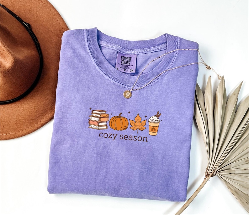May include: A lilac Comfort Colors t-shirt featuring a seasonal graphic. The design includes books, a pumpkin, a leaf, and a coffee cup. The words "cozy season" are printed below. A gold necklace and a brown hat are also visible in the image.