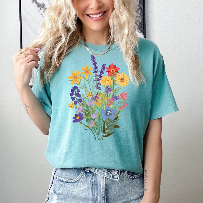 May include: A turquoise blue t-shirt with a colorful floral bouquet print. The flowers are in shades of yellow, orange, red, purple, and blue.