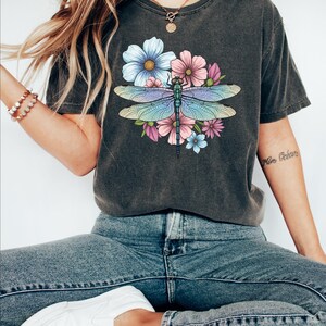 May include: Dark gray t-shirt featuring a colorful dragonfly design with blue, pink, and purple flowers. The dragonfly has iridescent wings. The shirt is paired with blue jeans and white sneakers.