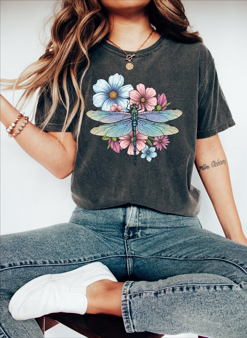May include: Dark gray t-shirt featuring a colorful dragonfly design with blue, pink, and purple flowers. The dragonfly has iridescent wings. The shirt is paired with blue jeans and white sneakers.