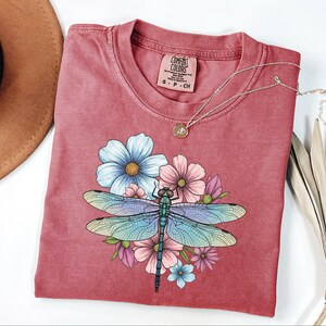 May include: A red-toned t-shirt featuring a colorful dragonfly design with blue, pink, and green hues, surrounded by floral accents. The shirt has a crew neck and short sleeves. A gold necklace with a pendant rests on the shirt.