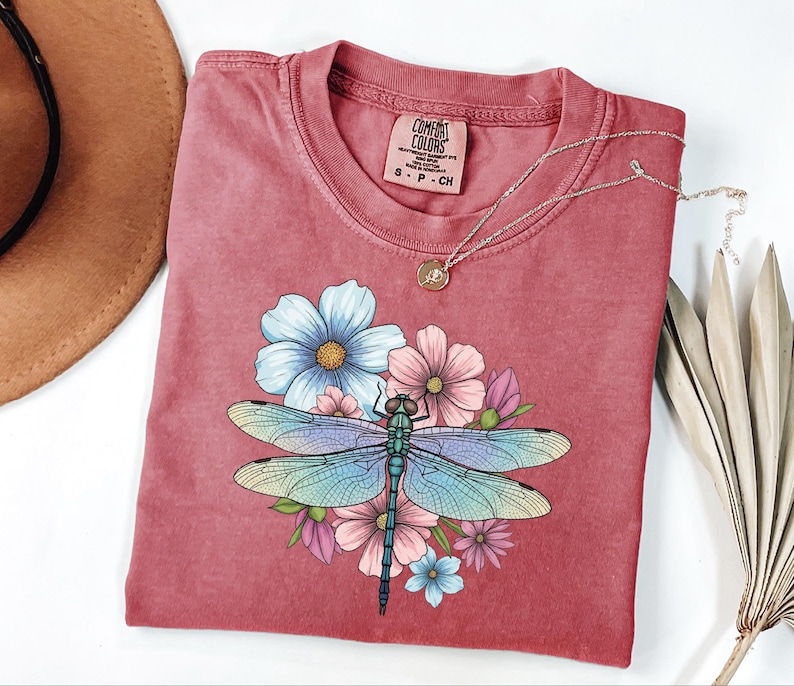 May include: A red-toned t-shirt featuring a colorful dragonfly design with blue, pink, and green hues, surrounded by floral accents. The shirt has a crew neck and short sleeves. A gold necklace with a pendant rests on the shirt.