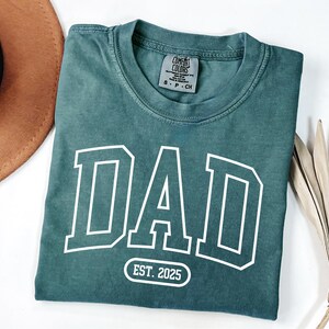 May include: A teal blue t-shirt with a white "DAD" graphic in a varsity style font. The shirt also has a white oval with the text "EST. 2025" printed on it.
