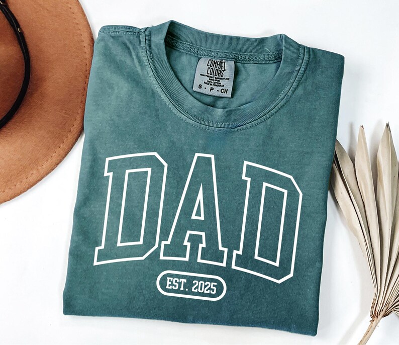 May include: A teal blue t-shirt with a white "DAD" graphic in a varsity style font. The shirt also has a white oval with the text "EST. 2025" printed on it.