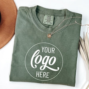 May include: A green t-shirt with a white circle that says "YOUR logo HERE" on it. The shirt is folded and laid flat on a white surface.