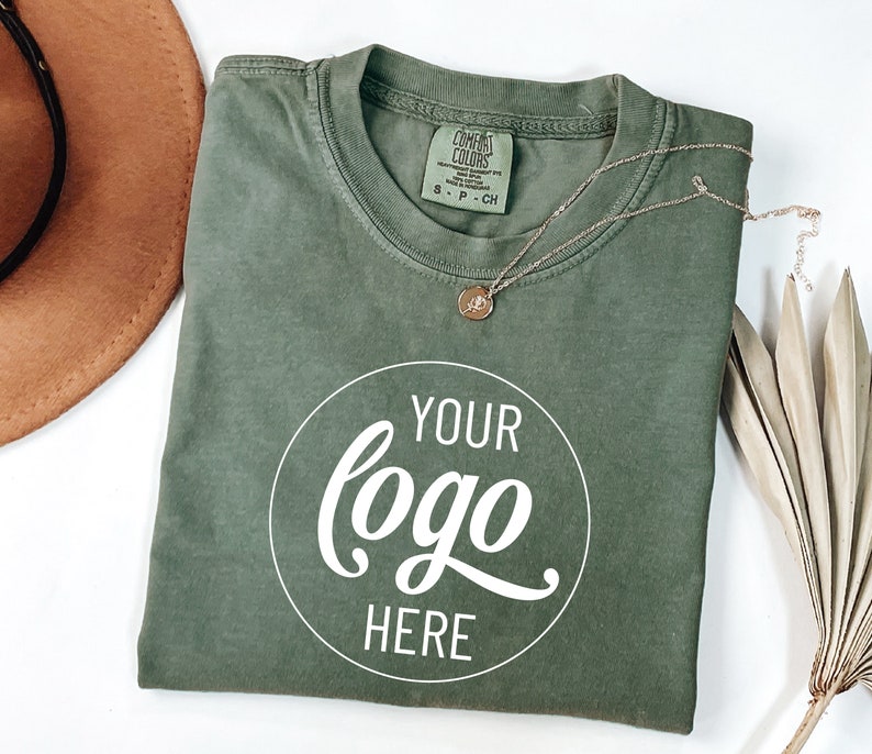 May include: A green t-shirt with a white circle that says "YOUR logo HERE" on it. The shirt is folded and laid flat on a white surface.