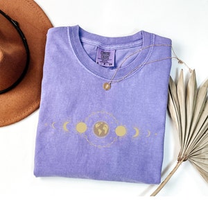 K&ouml;nnte beinhalten: Ein lilafarbenes T-Shirt mit einem goldenen, himmlischen Design, das Sonne, Mond und Erde zeigt. Das Shirt hat den Text "COMFORT COLORS" auf dem Etikett.