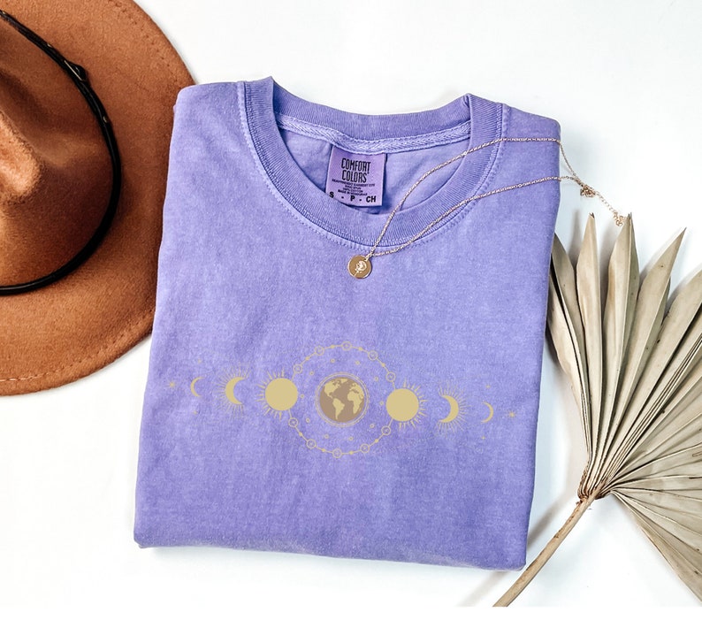 K&ouml;nnte beinhalten: Ein lilafarbenes T-Shirt mit einem goldenen, himmlischen Design, das Sonne, Mond und Erde zeigt. Das Shirt hat den Text "COMFORT COLORS" auf dem Etikett.