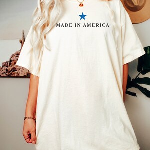 K&ouml;nnte beinhalten: Ein wei&szlig;es Oversize-T-Shirt mit einem blauen Stern und dem Aufdruck "MADE IN AMERICA" auf der Vorderseite.