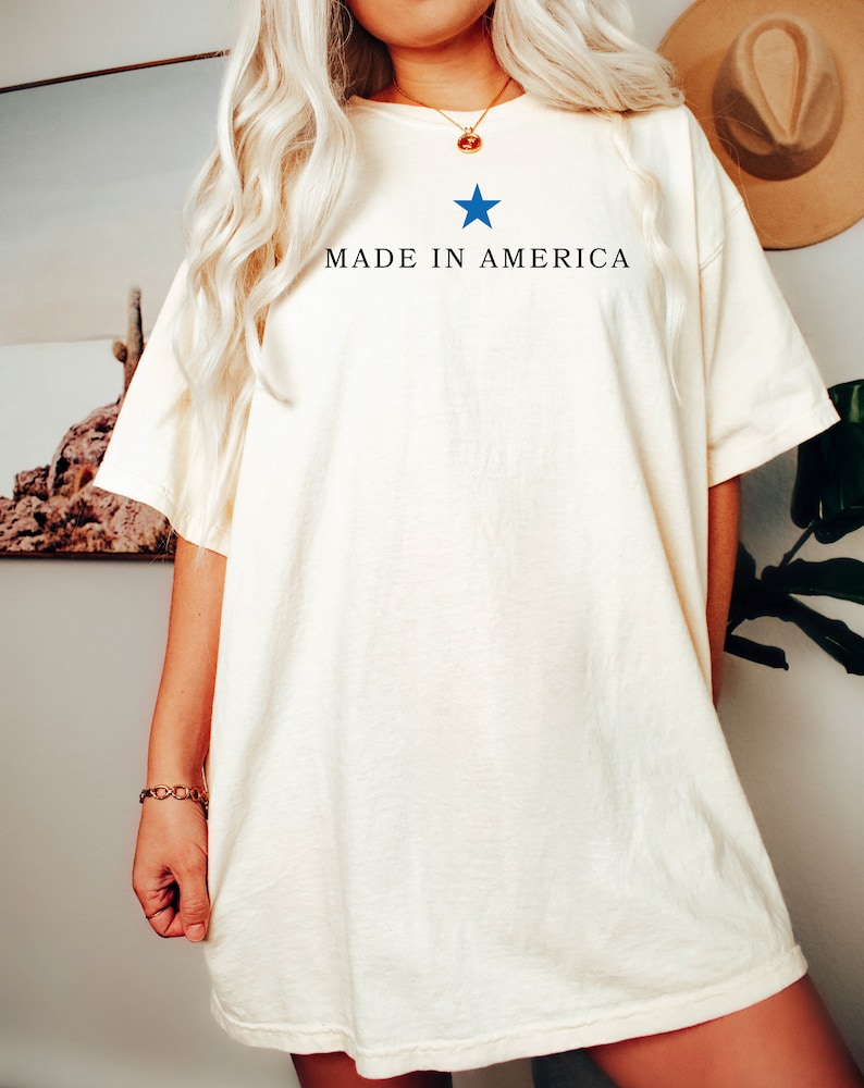 K&ouml;nnte beinhalten: Ein wei&szlig;es Oversize-T-Shirt mit einem blauen Stern und dem Aufdruck "MADE IN AMERICA" auf der Vorderseite.