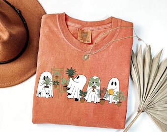 May include: A coral-colored t-shirt with a graphic of four ghosts holding potted plants. The text "Comfort Colors" is on a brown tag attached to the shirt.