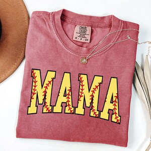May include: A red t-shirt with the word "MAMA" in yellow letters with red stitching around the letters, resembling a baseball. The shirt is folded and laid flat on a white surface.