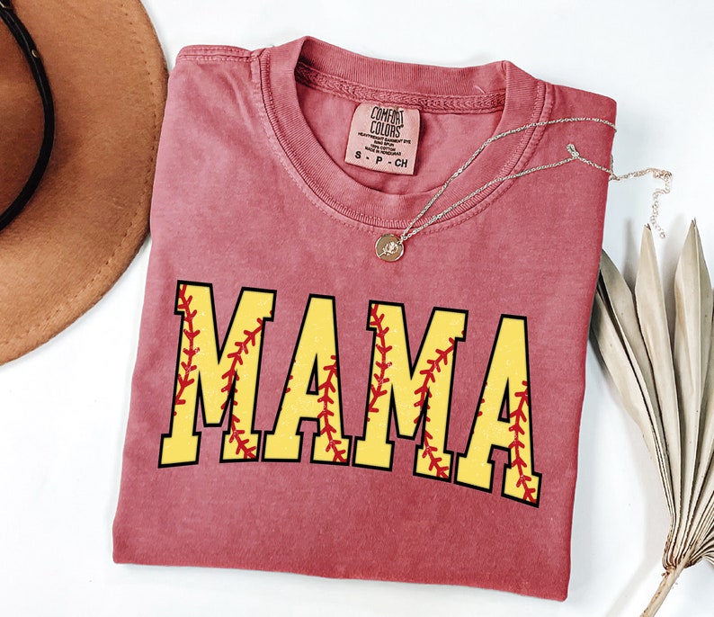 May include: A red t-shirt with the word "MAMA" in yellow letters with red stitching around the letters, resembling a baseball. The shirt is folded and laid flat on a white surface.