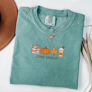 May include: A teal Comfort Colors t-shirt featuring a fall-themed graphic. The graphic includes a stack of books, a pumpkin, a leaf, and a coffee cup with the words "cozy season" below. A silver necklace with a pendant rests on the shirt.