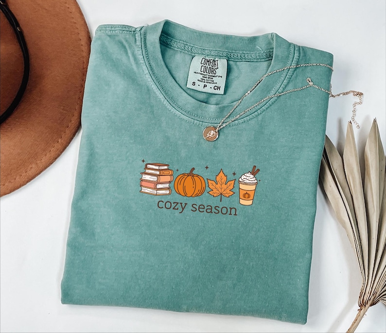May include: A teal Comfort Colors t-shirt featuring a fall-themed graphic. The graphic includes a stack of books, a pumpkin, a leaf, and a coffee cup with the words "cozy season" below. A silver necklace with a pendant rests on the shirt.