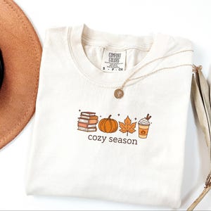May include: Off-white t-shirt featuring a seasonal graphic. The design includes a stack of books, a pumpkin, a maple leaf, and a coffee cup, with the words "cozy season" below. A gold necklace is draped across the shirt.