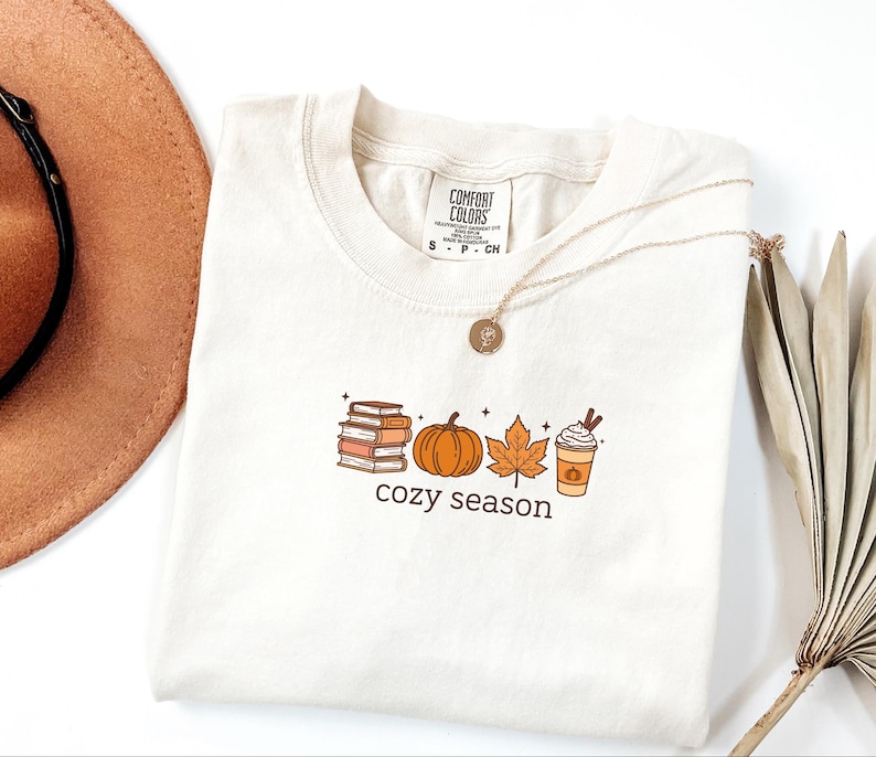 May include: Off-white t-shirt featuring a seasonal graphic. The design includes a stack of books, a pumpkin, a maple leaf, and a coffee cup, with the words "cozy season" below. A gold necklace is draped across the shirt.