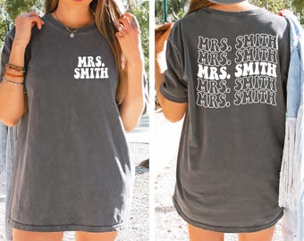 May include: Dark gray t-shirt with the text "MRS. SMITH" in white on the front and back. The back of the shirt repeats the text in a stacked design. The shirt has short sleeves and a relaxed fit.