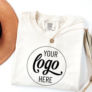 May include: A white t-shirt with a black circle that says "YOUR logo HERE" in a cursive font. The shirt is folded and laid flat on a white surface. A brown hat and a gold necklace are also visible in the image.