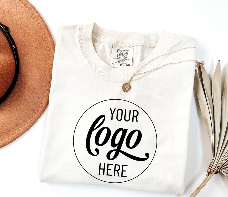May include: A white t-shirt with a black circle that says "YOUR logo HERE" in a cursive font. The shirt is folded and laid flat on a white surface. A brown hat and a gold necklace are also visible in the image.