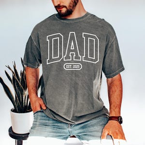 May include: A grey t-shirt with the word "DAD" in white outlined letters. The text "EST. 2025" is below the word "DAD".