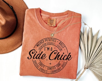 May include: A coral-colored t-shirt with a crew neck and the text "I'M A Side Chick" in a circular design. The design includes the words "Mashed Potatoes," "Rolls," "Stuffing," "Gravy," and "Candied Yams." A gold necklace and a brown hat are also in the image.