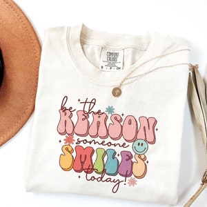 May include: A beige t-shirt with a retro-style graphic that says "be the reason someone smiles today!" in rainbow colors with flowers and a smiley face.