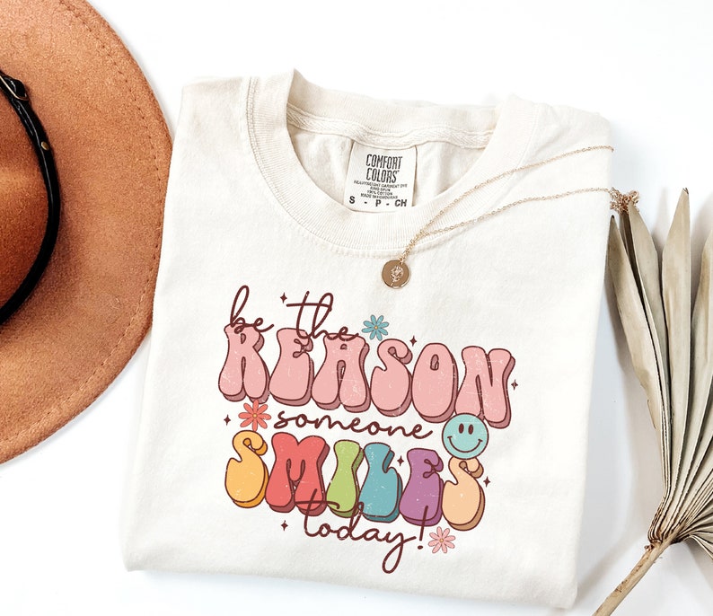 May include: A beige t-shirt with a retro-style graphic that says "be the reason someone smiles today!" in rainbow colors with flowers and a smiley face.