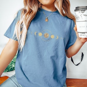 K&ouml;nnte beinhalten: Ein blaues T-Shirt mit einem Aufdruck einer stilisierten Erde, umgeben von Sonnen und Monden. Der Aufdruck ist in Gold und Gelb.